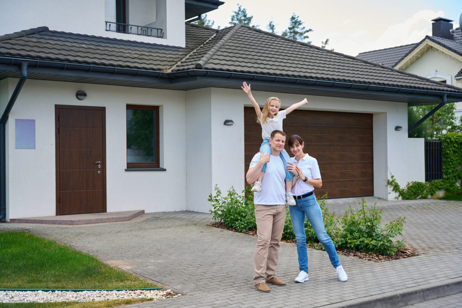 Pleased mother and daughter on father's shoulders in front of new townhouse Family smiling outside their modern home.